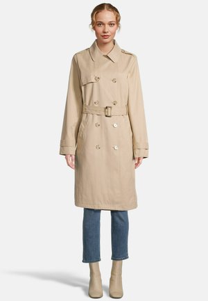 Vrouw staand, draag een beige dubbelgeknoopte trenchcoat met een riem in de taille, cropped blauwe jeans en beige enkellaarsjes.