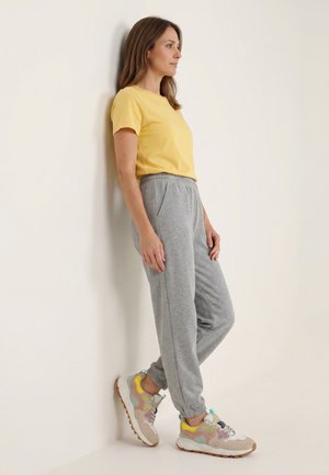 Frau lehnt an weißer Wand, trägt ein gelbes Kurzarmshirt, graue Jogginghose und bunte Sneaker mit pinken, gelben und türkisfarbenen Akzenten.