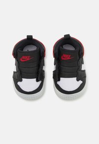 Sorte og hvite Nike-sneakers med en glatt skinnoverdel, borrelåsstropper og røde detaljer på hælen og logoen. Teksturert gummisåle.