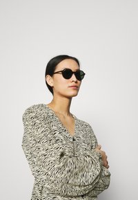 Occhiali da sole rotondi neri con lenti scure, indossati da un modello in una blusa a righe zebrate bianca e nera con scollatura a V.