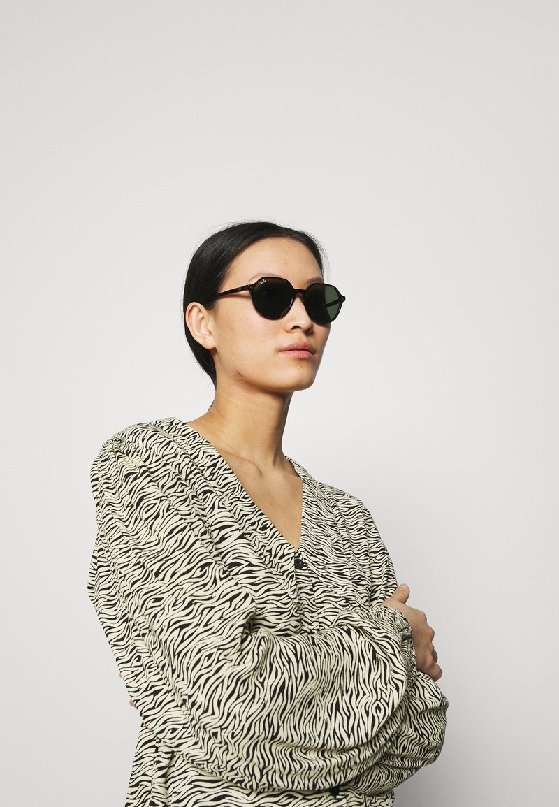 Occhiali da sole rotondi neri con lenti scure, indossati da un modello in una blusa a righe zebrate bianca e nera con scollatura a V.