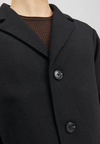 Svart ullmix-kappa med slitskrage, försedd med två svarta knappar och en mjuk yta, över en brun stickad tröja.