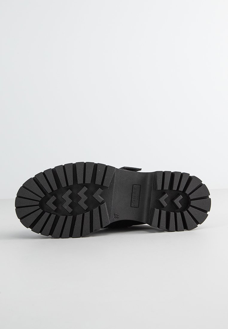 Semelle de botte en caoutchouc noir avec de profonds motifs de traction et un motif en chevron. Présente un marquage en relief et une section de orteil arrondie.