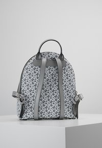 Grå mönstrad ryggsäck med två axelremmar och ett handtag på toppen. Har en texturerad utsida och en distinkt logodesign.