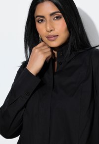 Kobieta o długich czarnych włosach, ubrana w czarną koszulę zapinaną na guziki, spoczywająca dłonią blisko brody na białym tle.