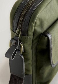 Grüne Canvas-Tasche mit schwarzen Lederelementen. Verfügt über einen Reißverschluss, Metallbeschläge und eine seitliche Tasche. Texturiertes Gewebe und glatte Oberfläche.