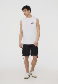 Canottiera bianca senza maniche con stampa "San Francisco", abbinata a pantaloni cargo neri con tasche laterali e sneakers bianche con dettagli grigi.