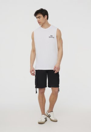 Canottiera bianca senza maniche con stampa "San Francisco", abbinata a pantaloni cargo neri con tasche laterali e sneakers bianche con dettagli grigi.