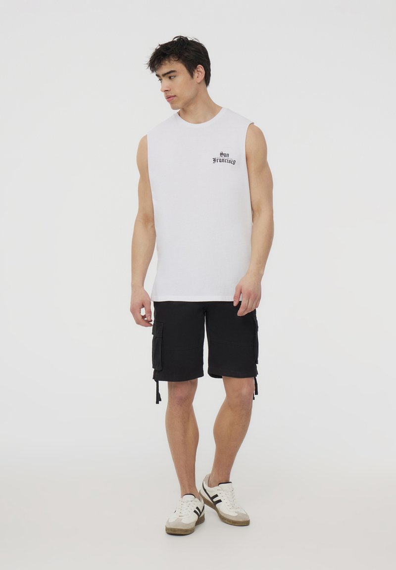 Canottiera bianca senza maniche con stampa "San Francisco", abbinata a pantaloni cargo neri con tasche laterali e sneakers bianche con dettagli grigi.