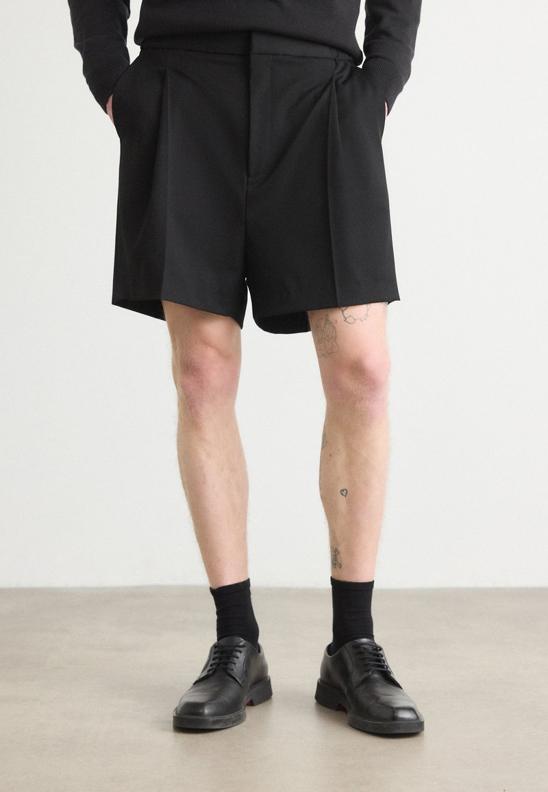 Schwarze maßgeschneiderte Shorts aus glattem Stoff, mit Vorderfalten und geradem Schnitt. Getragen mit schwarzen Socken und Schnürschuhen.