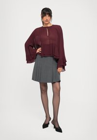 Blusa trasparente bordeaux con dettaglio a chiave, maniche fluide, abbinata a una gonna pieghettata grigia. La modella indossa tacchi a punta nera e collant trasparenti.