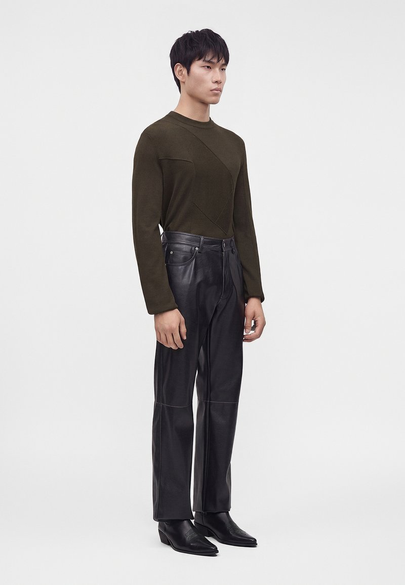 Young man stands facing slightly right wearing dark olive sweater, black leather pants, and black pointed dress shoes against a plain background.