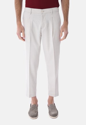Uomo con pantaloni plissettati color avorio sporco e mocassini in camoscio grigio, in piedi con le braccia rilassate lungo i fianchi contro uno sfondo semplice.