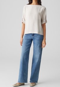 Helles beigefarbenes Oberteil aus Leinenmischung mit weitem Ausschnitt und umgeschlagenen kurzen Ärmeln, kombiniert mit hoch taillierten, weit geschnittenen blauen Jeans und beigen Ballerinas.