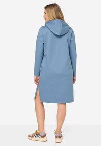 Robe bleue à capuche en tissu doux, avec des manches longues, des fentes sur les côtés et une coupe décontractée. Assortie à des baskets colorées.