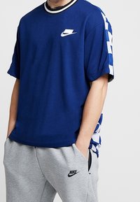 Námořnická modrá tričko s krátkým rukávem s bílým logem Nike na hrudi, s černobílým pruhovaným límcem a kostičkovanými modrými bočními akcenty.