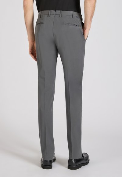 Uomo che indossa pantaloni eleganti grigi aderenti e scarpe nere, mostrato di spalle con le mani in tasca su sfondo semplice.