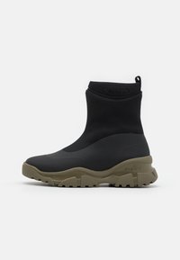 Botas altas negras con un acabado suave y mate. Suela de goma en verde oliva, con un agarre texturizado. Logo estampado en el talón.