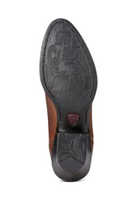 Zapato de cuero marrón con una suela de goma negra con patrón. Los detalles notables incluyen un emblema burdeos y un diseño de punta afilada y elegante.