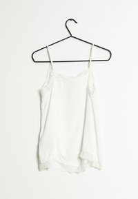 Vit linne med tunna band, med spetsdekoration längs halsringningen och nederkant. Tillverkad av mjukt, lätt tyg. Hänger på en svart krok.