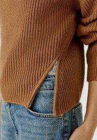 Bruine gebreide trui met een diagonale zoom en een zilveren ritsdetail, gecombineerd met lichtblauwe denim jeans. Geverfd materiaal.