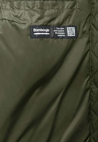 Interno di una giacca trapuntata verde oliva con etichetta nera che riporta la scritta "Bomboogie" contenente un codice QR e testo per informazioni sul prodotto. Texture liscia e lucida.