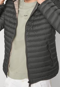 Person som bär en delvis nedknäppt svart vadderad jacka över en ljusgrön rundhalsad tröja och vita byxor, och justerar jackans krage.