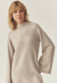 Beige kötött pulóver, magas nyakú, hosszú ujjú és puha tapintású. A design laza fazonú, bordás mandzsettákkal.