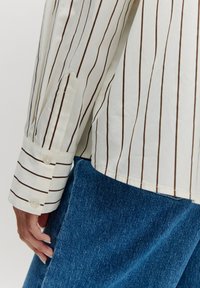 Weißes Hemd mit schmalen braunen Streifen, mit einem knöpfbaren Manschette. Kombiniert mit blauen Denim, die einen strukturierten Stoff zeigen.