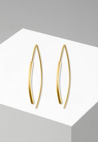 Gullfargede metalløreringer med en buet, avlang form. Glatt tekstur med subtilt skinnende finish. Minimalistisk design på en hvit utstilling.