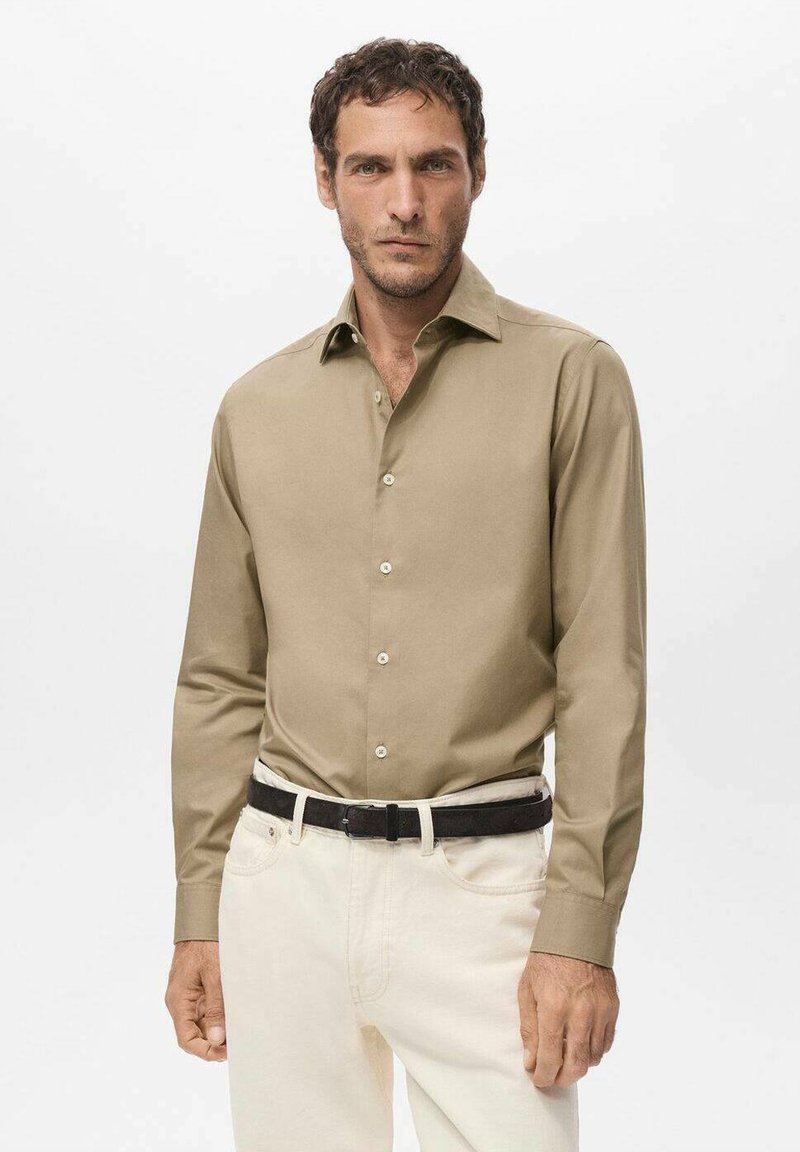 Camisa de botones de manga larga en color beige, hecha de una tela suave, con un cuello clásico y botones en contraste, combinada con pantalones de color claro y un cinturón negro.