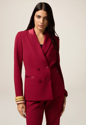 Femme portant un blazer bordeaux à double boutonnage sur mesure et un pantalon assorti, accessoirisée de bracelets beige et rose à son poignet.