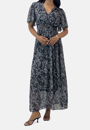 Kvinde iført en sort-hvid blomsterprintet maxikjole med pufærmer, v-hals og sorte stropper, stående foran en ensfarvet baggrund.