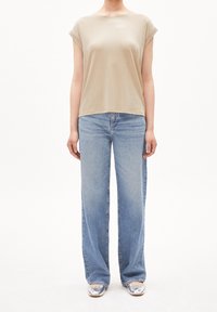 Camiseta beige de manga corta y cuello redondo combinada con unos jeans de pata ancha en azul claro. El modelo lleva unas bailarinas plateadas brillantes. Corte sencillo y relajado.