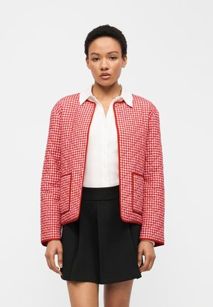 Γυναίκα που φοράει κόκκινο και λευκό καρό καπιτονέ μπουφάν πάνω από μια λευκή μπλούζα και μαύρη πλισέ φούστα, στέκεται μπροστά σε απλό φόντο.