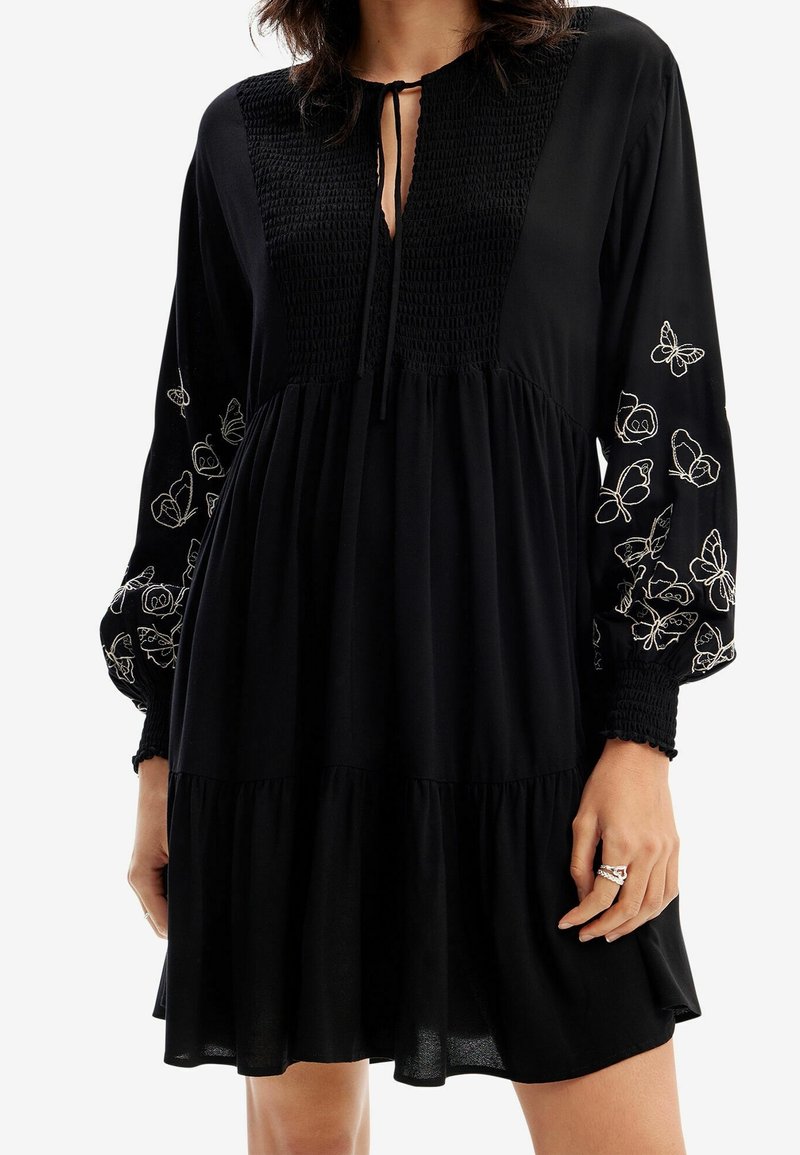 Femme portant une robe noire avec corsage smocké, col à nouer et manches longues brodées de motifs de papillons blancs.