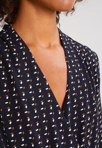 Haut enveloppant bleu marine foncé avec un décolleté en V profond, présentant un motif de petites formes géométriques blanches et dorées. Tissu lisse avec un léger éclat.