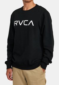 Homme portant un sweatshirt noir à col rond avec le logo blanc "RVCA" et un pantalon beige, se tenant avec une main dans la poche.
