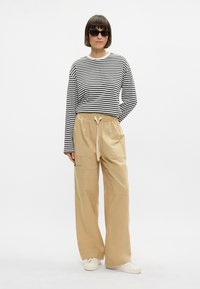 Μαύρο και λευκό ριγέ πουκάμισο με μακριά μανίκια συνδυασμένο με μπεζ παντελόνι cargo σε φαρδιά γραμμή. Τα παντελόνια διαθέτουν τσέπες; το μοντέλο φοράει μαύρα γυαλιά ηλίου και λευκά αθλητικά παπούτσια.