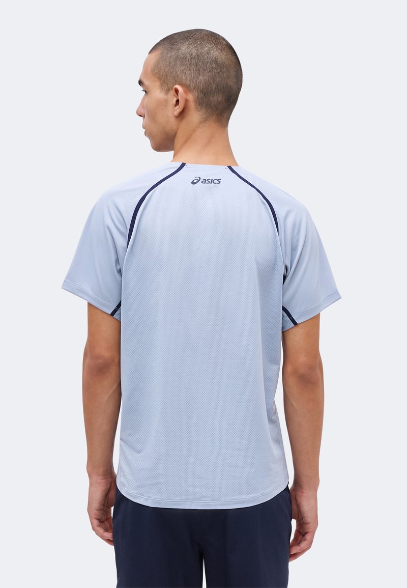 Mand med kort hår iført en lysegrå Asics atletik T-shirt og mørke shorts, set bagfra mod en ensartet baggrund.
