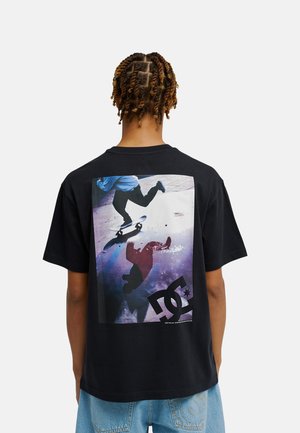Osoba z zaplecionymi włosami ma na sobie czarny T-shirt z kolorową grafiką deskorolki i logo DC Shoes na plecach, zestawiony z niebieskimi dżinsami.