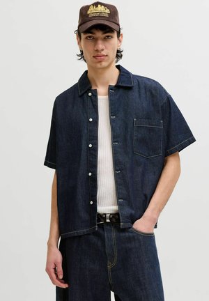 Jeune homme portant une chemise en jean sombre à manches courtes boutonnée sur un top en maille blanc, un jean foncé, une ceinture tissée et une casquette marron avec une broderie jaune.