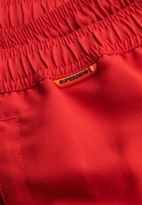 Rode zwembroek met een elastische tailleband, voorzien van een zwart en oranje "SUPERDRY" logo-label. Gladde textuur en omgevouwen ontwerp.