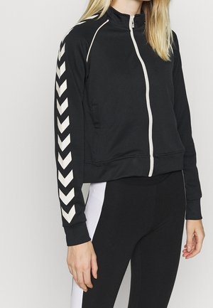Sort zip-jakke med høj krave, hvid chevron-stribe på ærmerne, lavet af blødt materiale. Har sidelommer og en tætsiddende design.