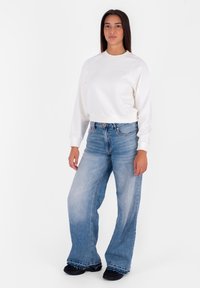 Felpa bianca corta con collo a giro e maniche lunghe, abbinata a jeans a gamba larga blu chiaro e scarpe nere. Stile semplice e casual.