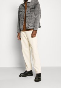 Giacca di denim grigia con colletto peloso, camicia marrone sotto, pantaloni in velluto a coste beige e stivaletti neri alla caviglia. Dettagli notevoli su tasche e bottoni.