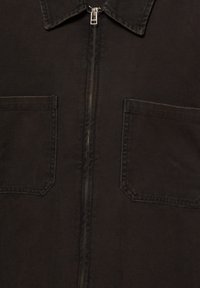 Veste en tissu foncé à l'avant avec une fermeture éclair en métal centrée entre deux poches carrées sur la poitrine et un col pointu.