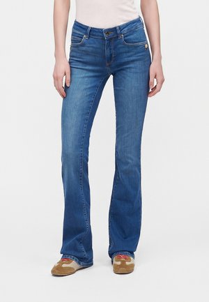 Kvinde iført blå bootcut-jeans og beige sneakers med røde snørebånd, stående mod en ensfarvet lys baggrund.