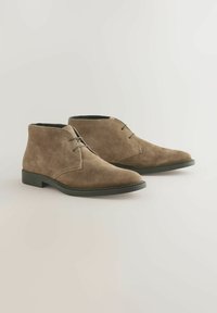 Brune ruskinds ankelstøvler med en rund tå, minimal syning og sort gummisål. Har et enkelt snørebåndsdesign til lukning.
