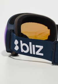 Snehové okuliare s jantárovým sklom a námorníckym látkovým páskom, ktorý obsahuje bielu značku "bliz", navrhnuté na pohodlie a výkon.